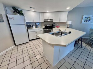 A modern kitchen with white cabinets, a central island with a sink, a microwave, a stove, and a refrigerator. - Gulf Shores Getaway! 2BR Pool Walk to Beach Pet Friendly (Gulf Shores)