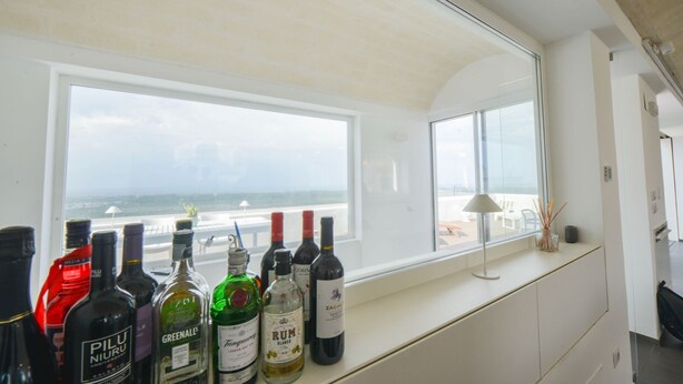 Ein Zimmer mit großem Fenster und Meerblick, einer Minibar mit verschiedenen Flaschen sowie einer Lampe am Fensterbrett.