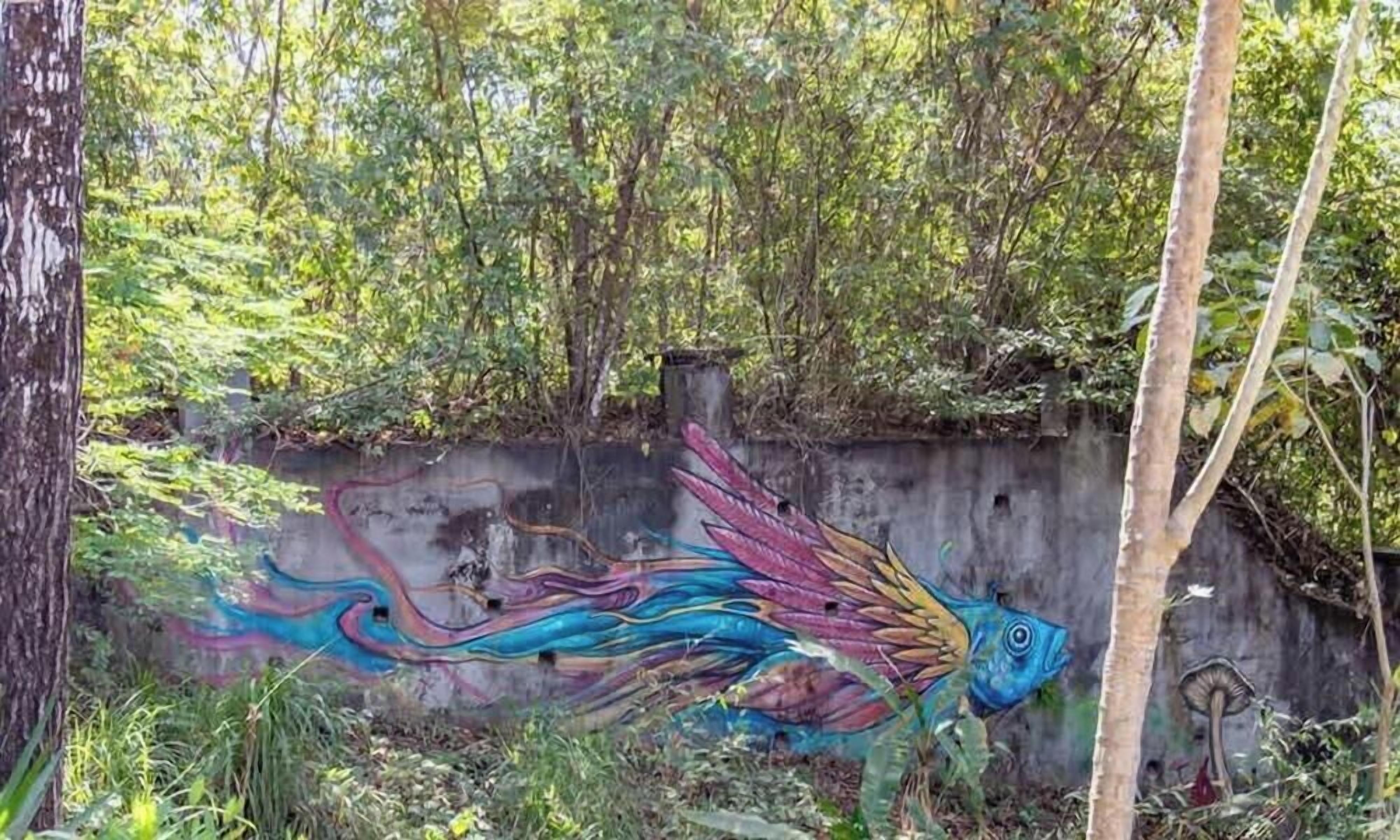 A mural of a colorful fish on a concrete wall surrounded by greenery.