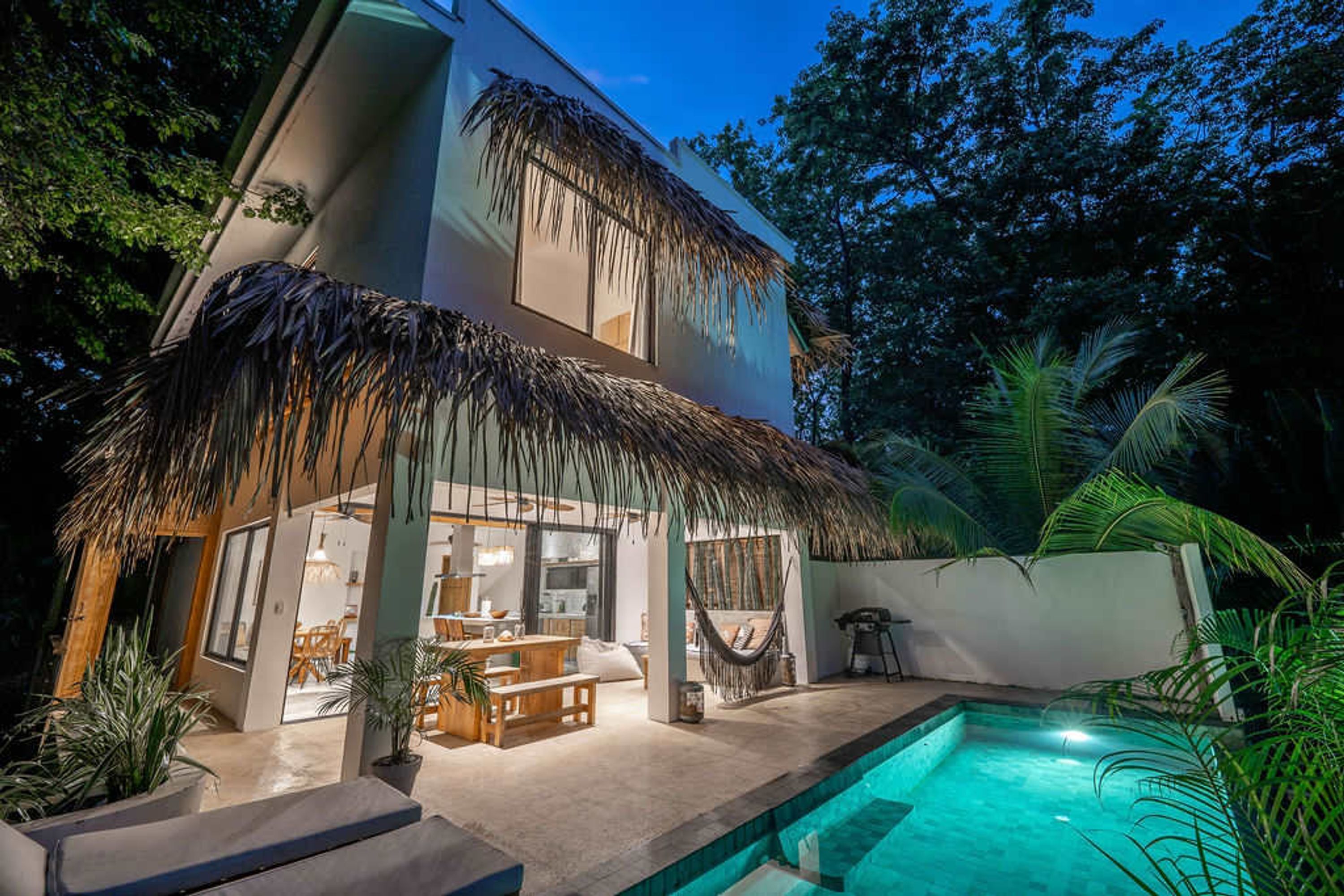 Una casa de dos pisos con piscina, mobiliario exterior y plantas tropicales.