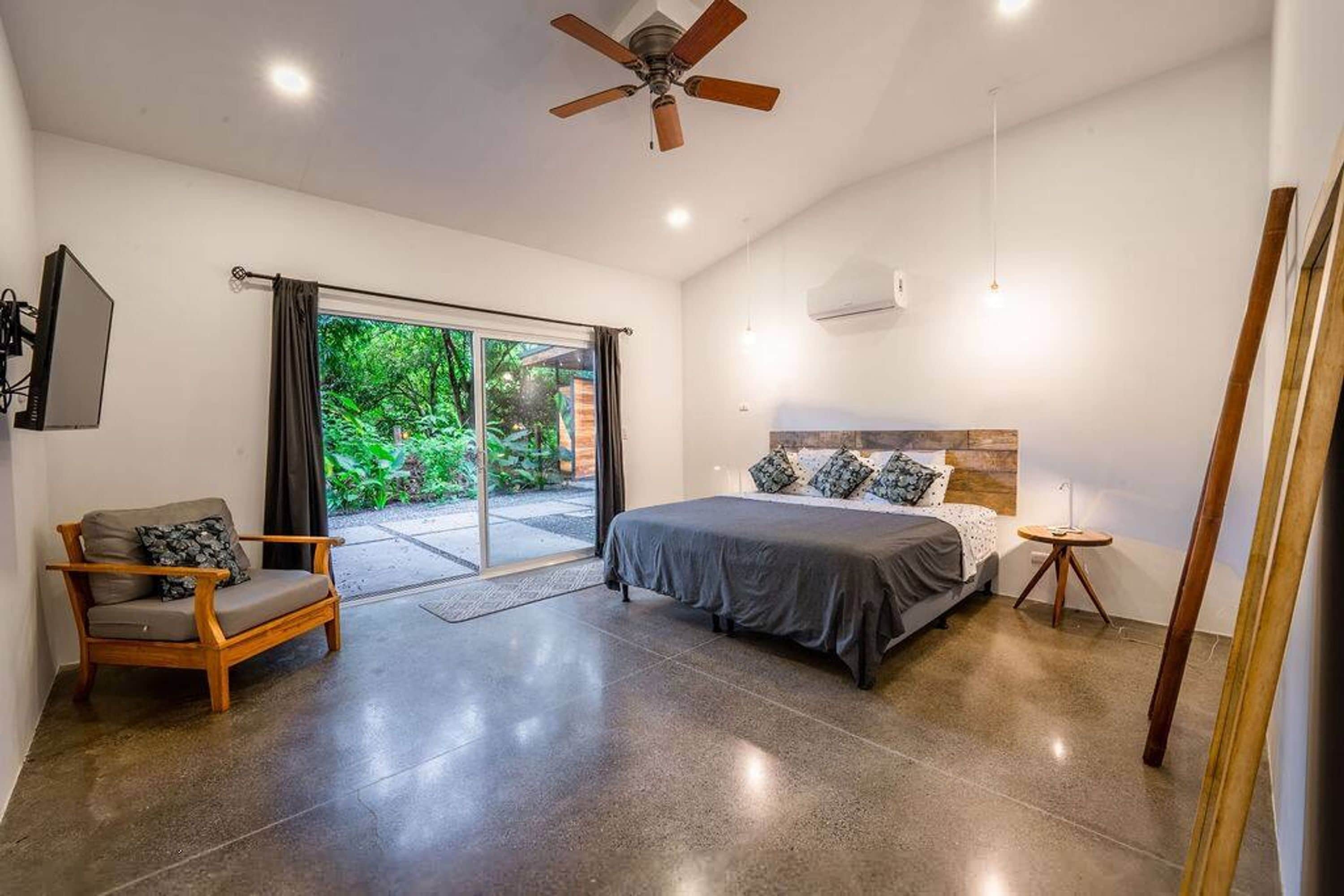 Un dormitorio con cama, una silla, ventilador de techo y vista a la vegetación por la ventana.