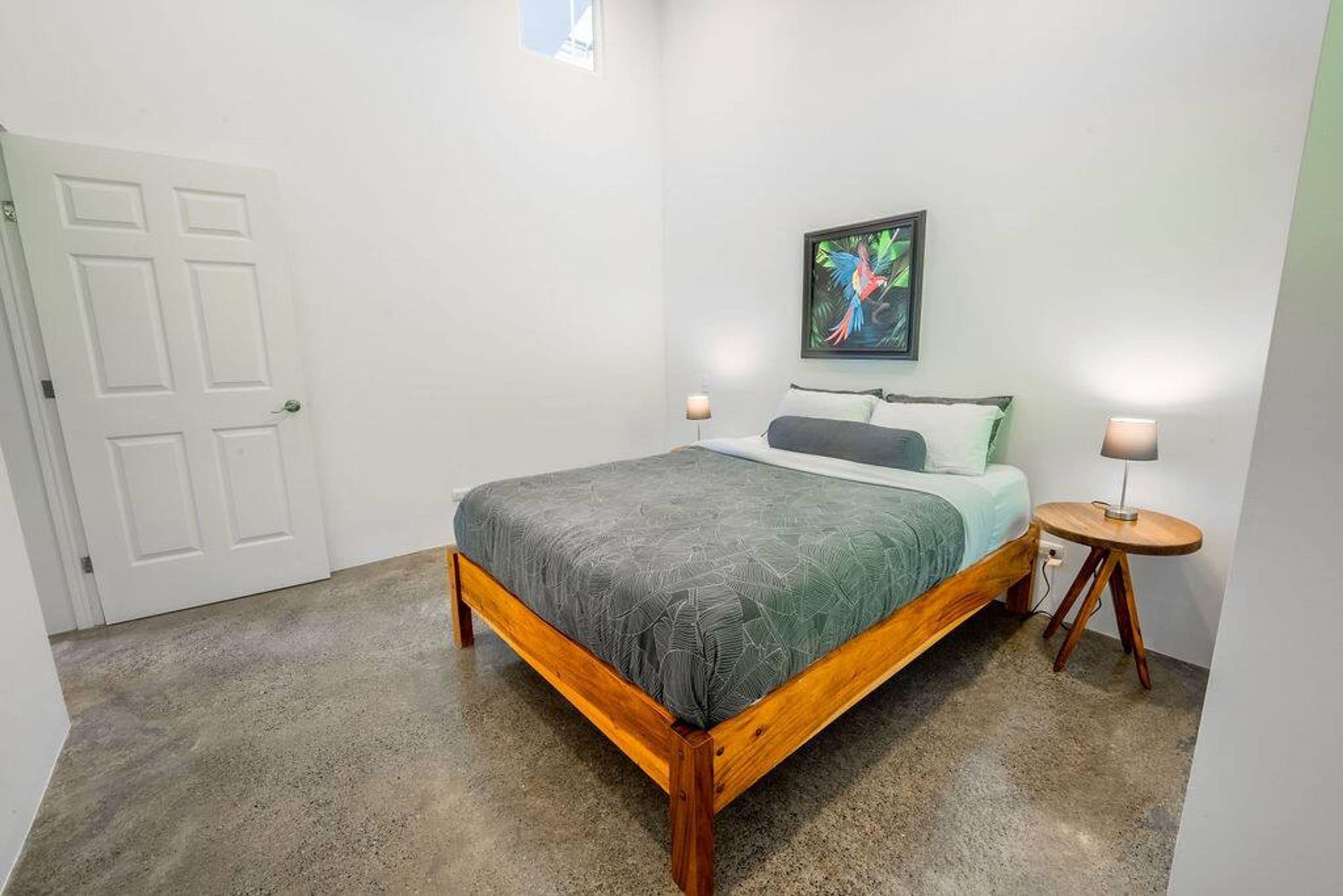 Un dormitorio con cama, una puerta, un cuadro colgado en la pared y una mesita de noche con lámpara.