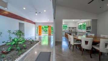 A modern kitchen with white cabinets, a dining area with a wooden table and white chairs, and a view of greenery through a glass door.