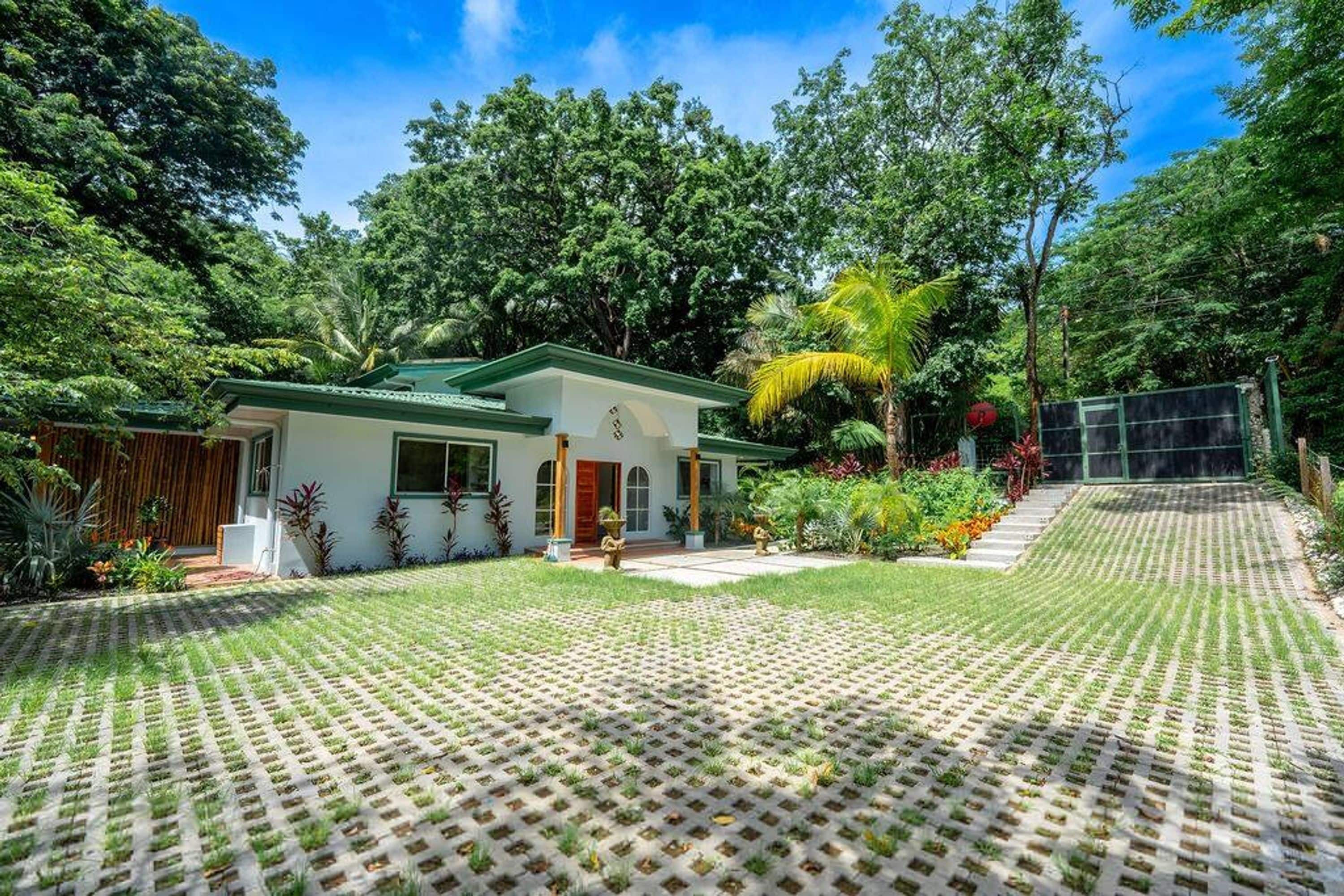 Una casa con techo verde, rodeada de abundante vegetación y un área pavimentada.