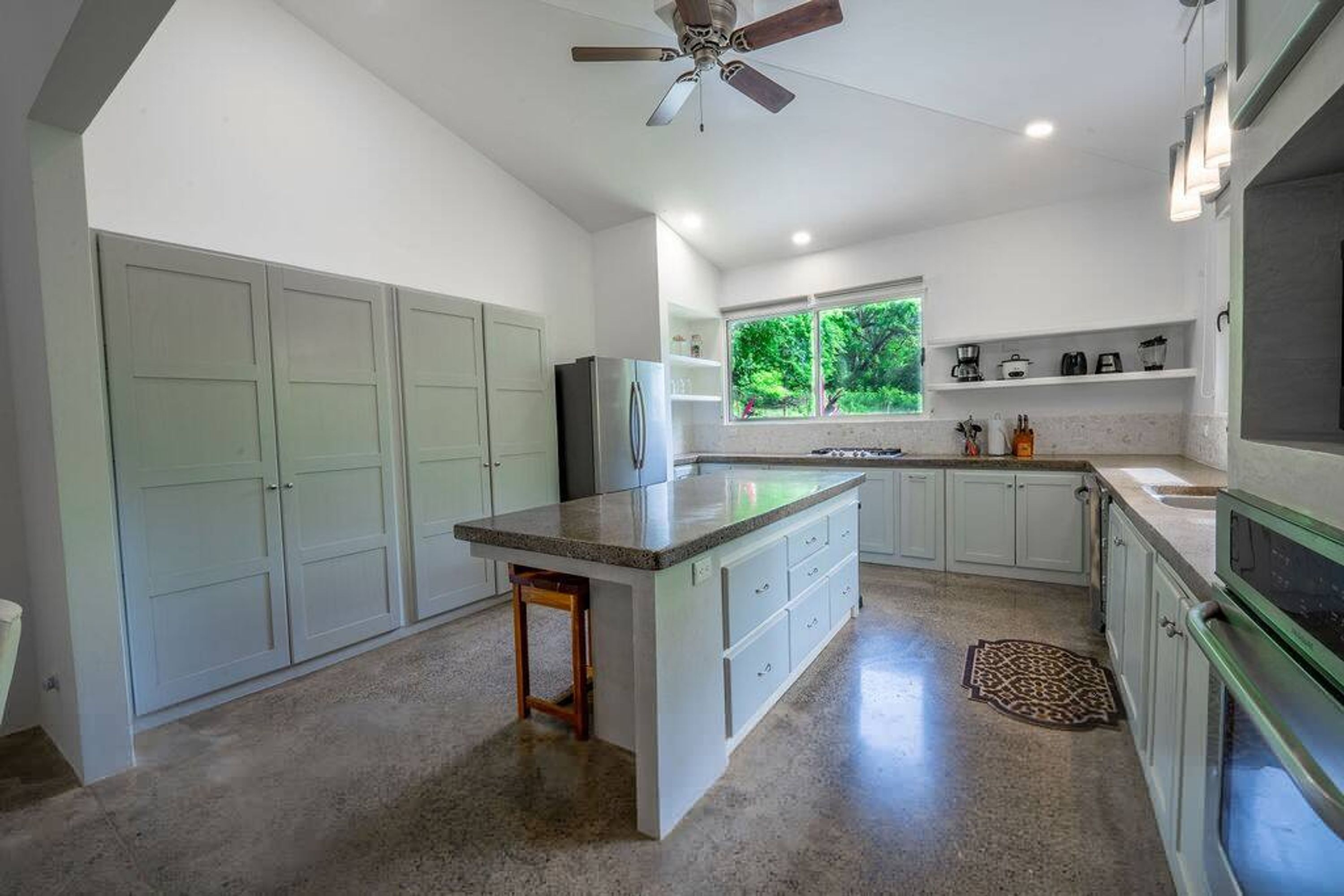A modern kitchen with a central island, white cabinetry, a large window, and a ceiling fan.