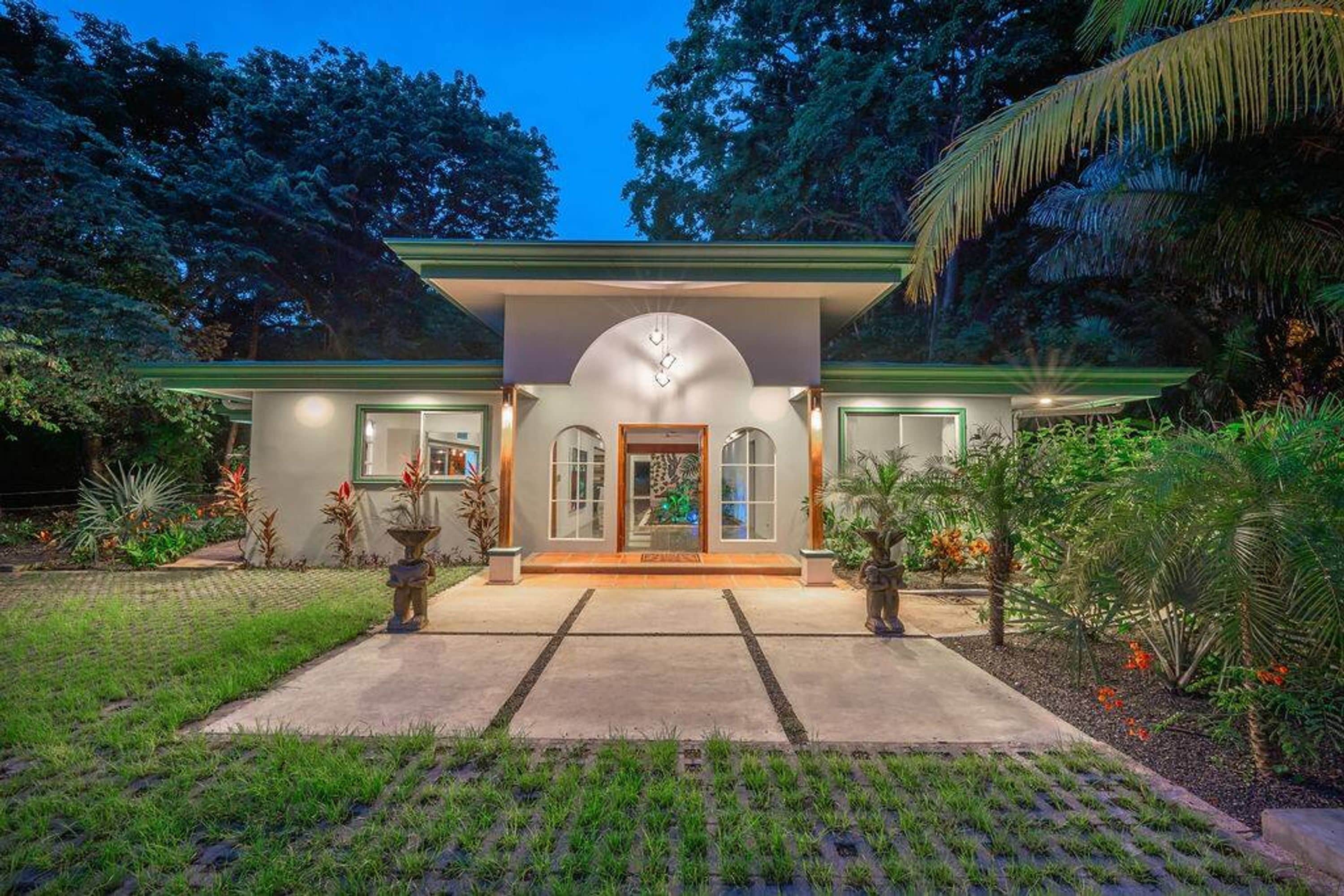 Una casa de dos pisos con fachada simétrica, una entrada central y un jardín cuidado con plantas tropicales.