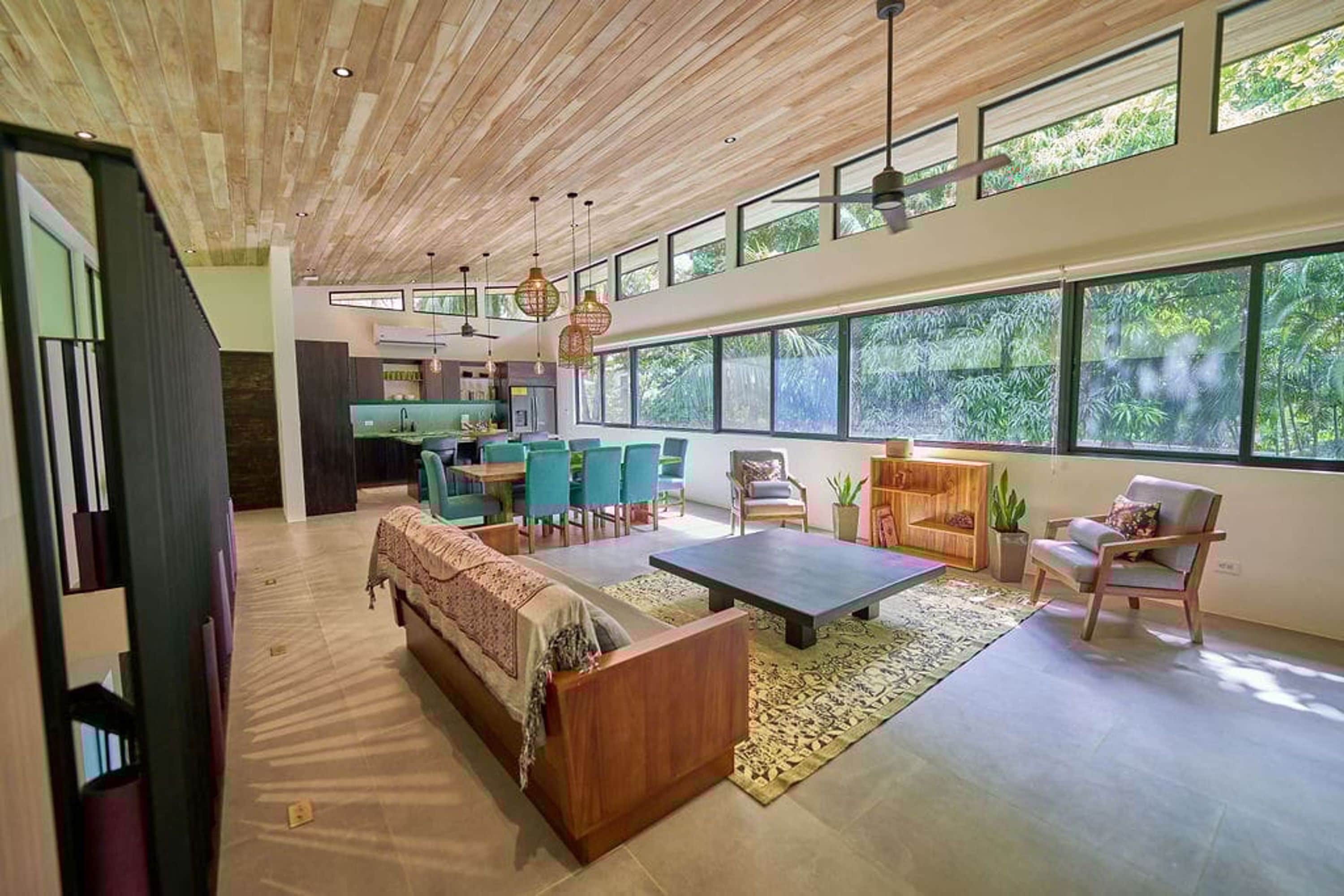 A spacious living area with a wooden ceiling, large windows, and a modern kitchen in the background.