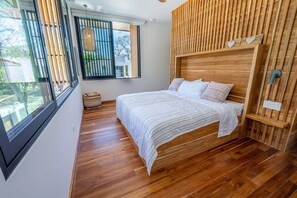 A bedroom with a wooden headboard, a bed with white bedding, a window with blinds, and a small round rug. - Brand New Casa Marea Walk to Beach (Nosara)