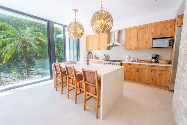 Cozinha moderna com armários de madeira, uma ilha central e uma grande janela com vista para um jardim tropical.