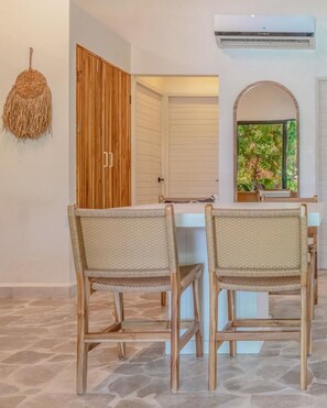 A room with a dining table, two wicker chairs, a woven wall hanging, a wooden door, and an air conditioning unit. - Casa Carlita at Hidden Palms (Nosara)