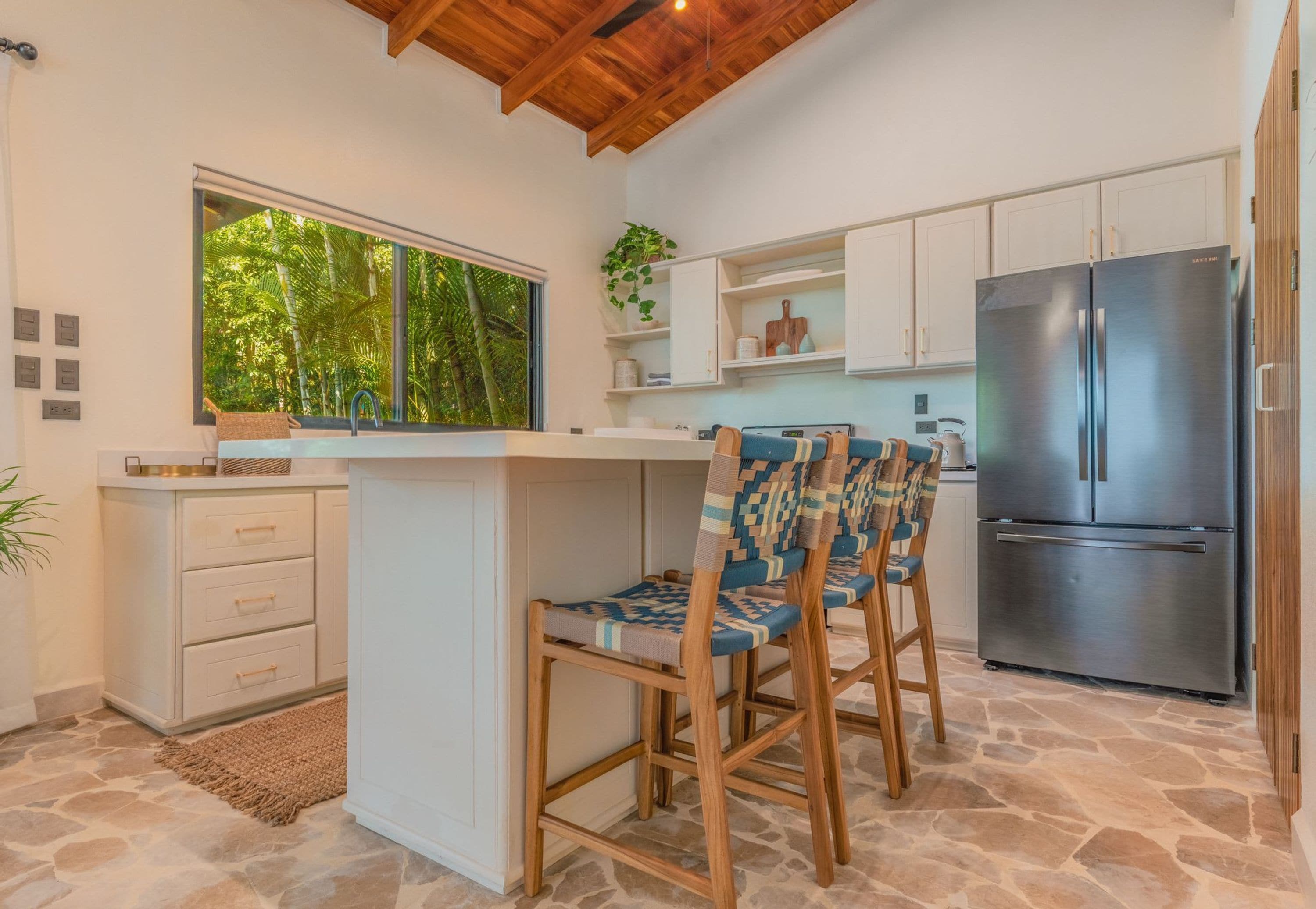 Una cocina con suelo de piedra, sillas de madera, un refrigerador de acero inoxidable y una ventana con vistas a zonas verdes.
