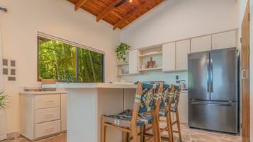 Una cocina con suelo de piedra, sillas de madera, un refrigerador de acero inoxidable y una ventana con vistas a zonas verdes.