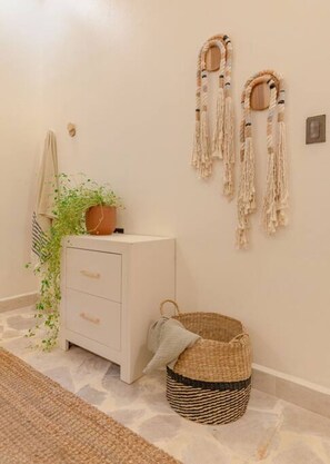 Une pièce au décor minimaliste, avec une commode blanche, un panier en osier, des décorations murales en macramé et une plante en pot.