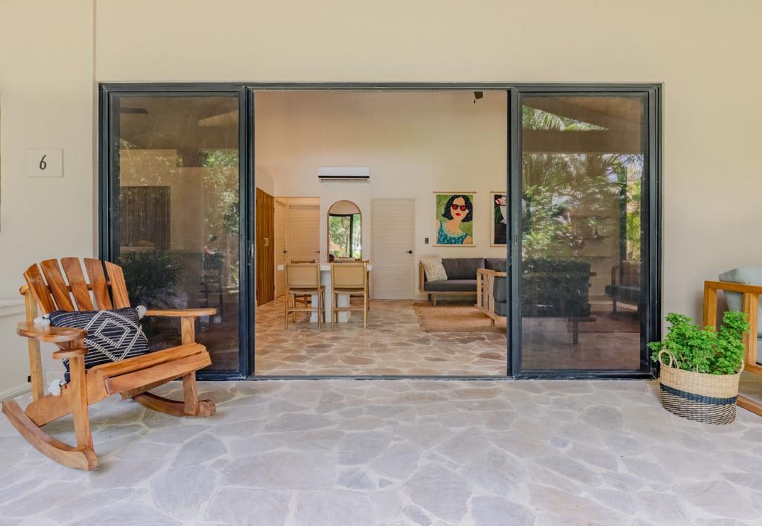 Um quarto com soalho de pedra, uma cadeira de balanço de madeira, uma mesa de jantar com cadeiras e uma porta de vidro deslizante que dá para outro quarto.