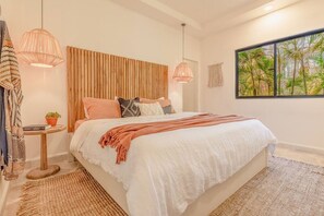 Une chambre avec un grand lit, une tête de lit en bois, deux lampes suspendues, une fenêtre donnant sur des palmiers et un tapis tissé.