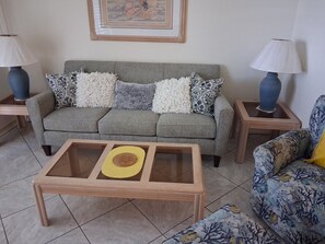 Ein Wohnzimmer mit einem grauen Sofa, gemusterten Kissen, einem Holztisch mit gelbem Untersetzer und zwei Nachttischen mit Lampen.
