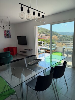 Un salon moderne avec une table en verre, des chaises, un canapé rouge, un téléviseur à écran plat et un balcon offrant une vue sur les collines.