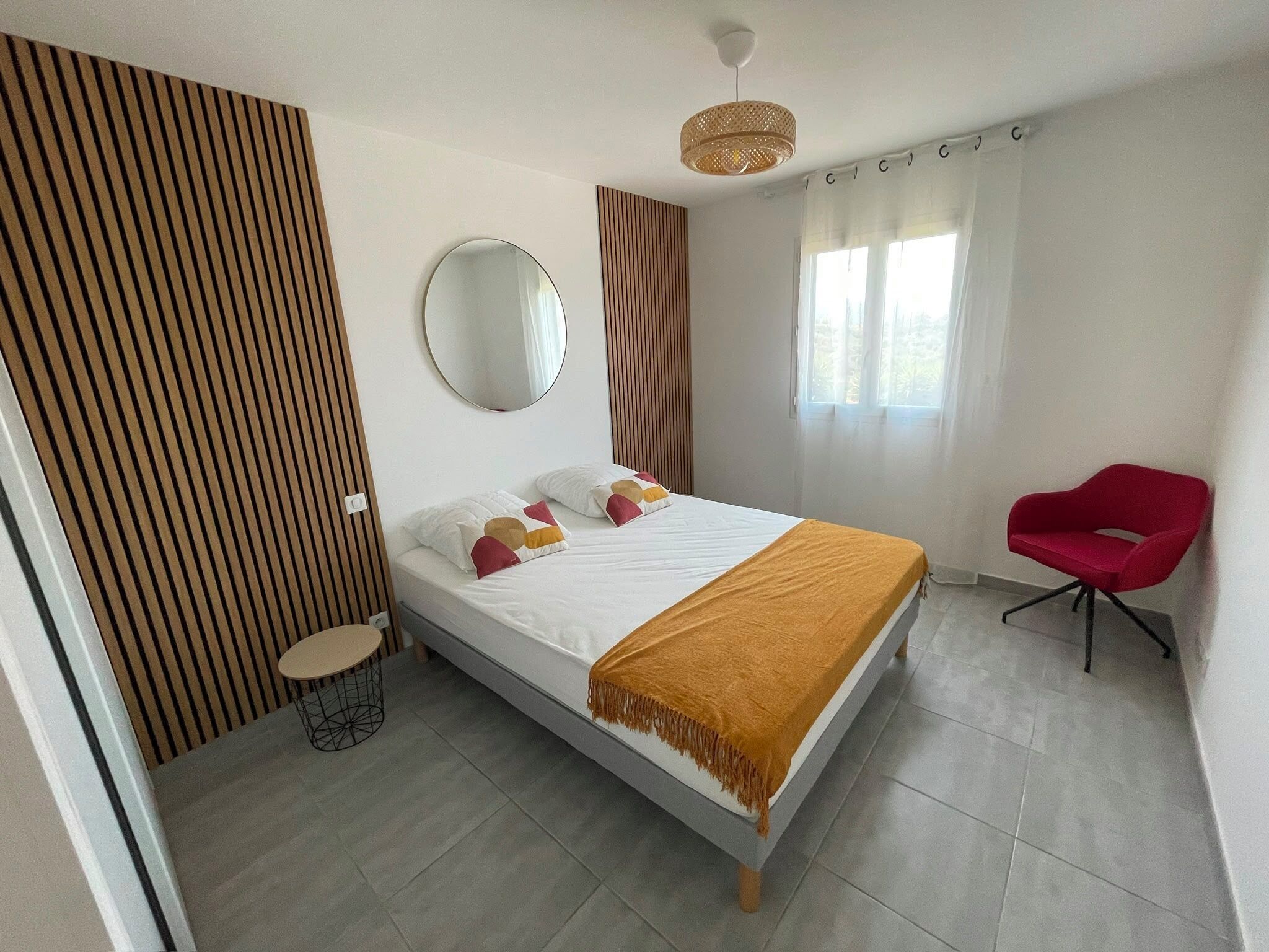 Une chambre à coucher avec un lit, un miroir rond, une chaise rouge et une fenêtre avec des rideaux.
