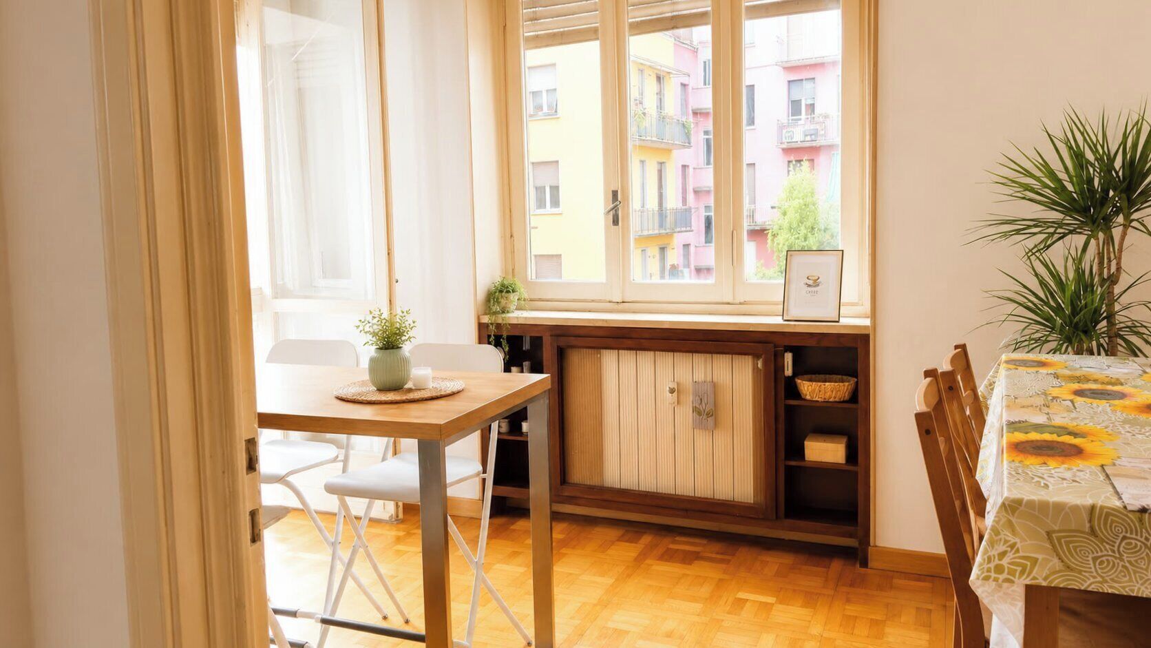 A dining area with a wooden table, chairs, a cabinet, and a window with a view of colorful buildings.