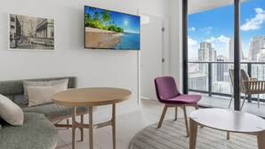 Ein modernes Wohnzimmer mit einer Couch, einem runden Tisch und einem Sessel. Ein großes Fenster bietet Blick auf die Stadtlandschaft.