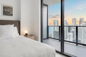 A bedroom with a large bed, a bedside table with a lamp, and a view of a cityscape through a large window. - 33rd-Floor City Retreat with Balcony Views in Miami G3308 (Miami Beach)