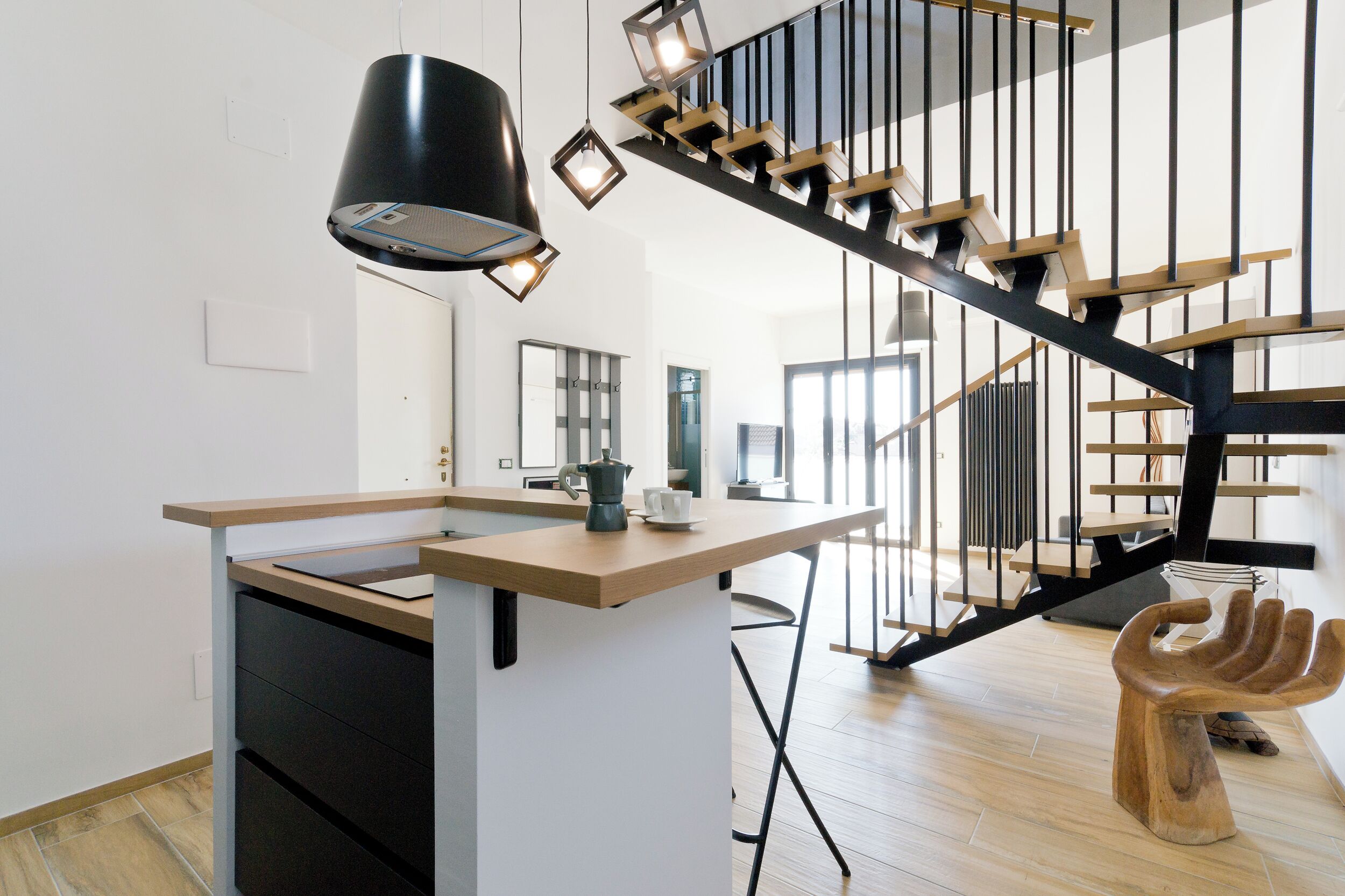 Une cuisine moderne dotée d’un îlot central, d’un escalier au design original et d’un tabouret en bois dont la forme rappelle celle d’une main.