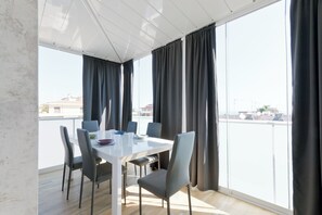 A modern dining area with a white table and grey chairs, large windows with dark curtains, and a view of buildings in the distance. - Pescara Beach Alba - One Bedroom Apartment, Sleeps 4 (Pescara)