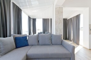 A modern living room with a grey sofa, multiple cushions, and large windows with grey curtains. - Pescara Beach Alba - One Bedroom Apartment, Sleeps 4 (Pescara)