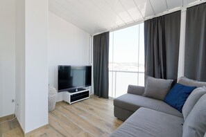 Ein modernes Wohnzimmer mit Flachbildfernseher, grauem Sofa und großen Fenstern mit Vorhängen.