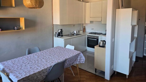 Una cocina compacta con armarios blancos, una mesa de comedor con mantel estampado y una lámpara colgante.