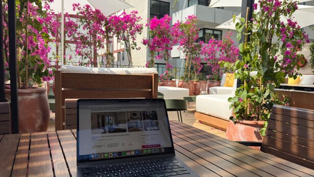 Ein Laptop auf einem Holztisch mit Blick auf Sitzgelegenheiten im Freien und Kübelpflanzen.