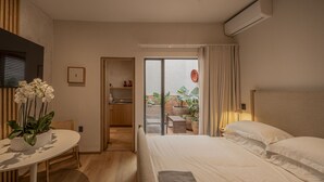 Ein Hotelzimmer mit einem Bett, einem kleinen Tisch mit Pflanze, einer Küchenzeile und einer Schiebetür zu einem Balkon mit Pflanzen.