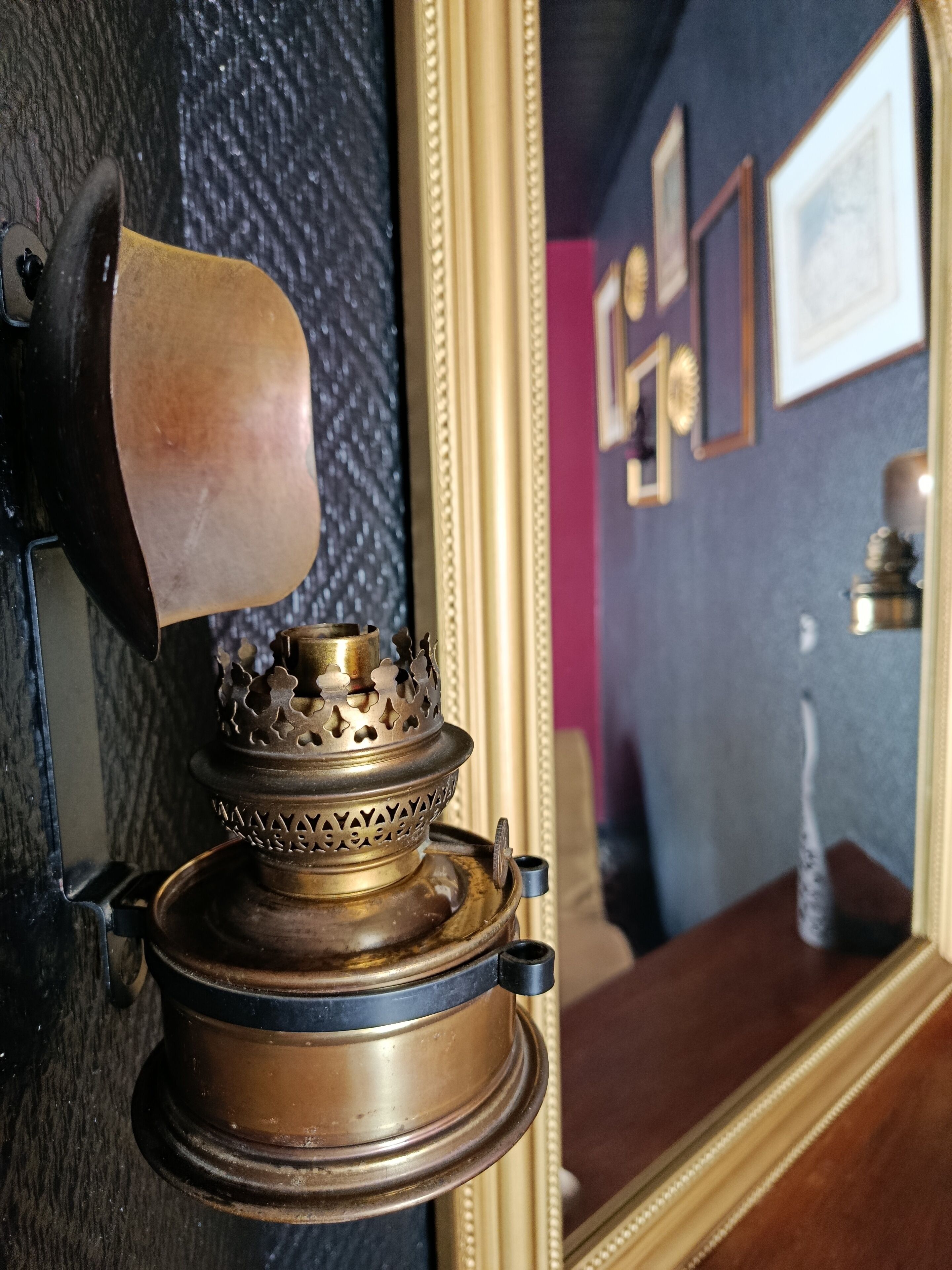 A brass lamp with intricate designs, a dark textured wall, and a mirror reflecting a framed picture.