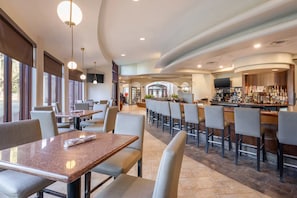 Ein modernes Restaurant mit Barbereich, das eine Mischung aus Sitzgelegenheiten bietet, darunter Barhocker und Tische, sowie eine Theke mit einer Auswahl an Flaschen.