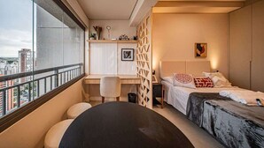 A modern bedroom with a bed, bedside table, and a desk with a chair. There is a window with a view of buildings.