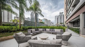 An outdoor seating area with a circular table and cushioned chairs, surrounded by greenery and tall buildings.