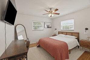 A bedroom with a bed, a desk, a television, a ceiling fan, and two windows with blinds. - The Birdhouse - Mid-Century Charm with a Speakeasy Lounge (Grove City)