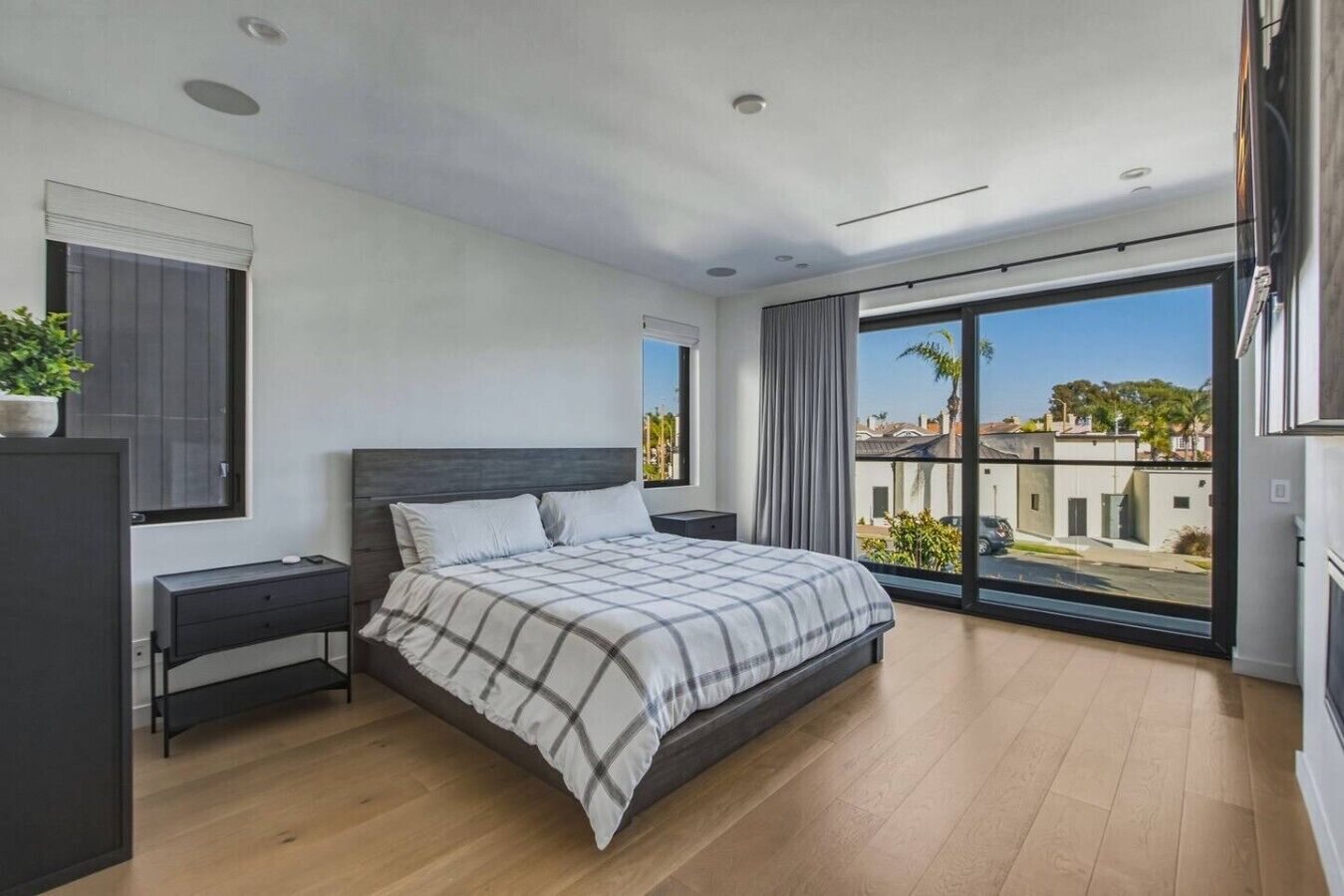 A modern bedroom with a large bed, a bedside table, a wardrobe, and a balcony with a view of buildings and palm trees.