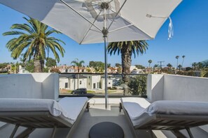 A balcony with two lounge chairs, a large umbrella, and a view of palm trees and buildings. - Luxury Home On 7th - Patio, Deck, Fire Pit (Huntington Beach)
