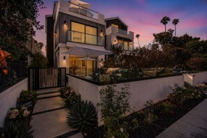 A modern two-story house with a balcony, surrounded by landscaped gardens and palm trees. - Luxury Home On 7th - Patio, Deck, Fire Pit (Huntington Beach)