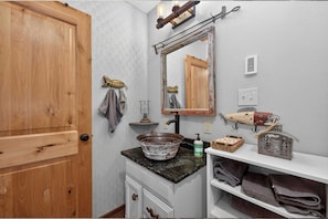 Uma casa de banho com porta de madeira, espelho, lavatório com cuba sobreposta, toalheiro e prateleira com toalhas.