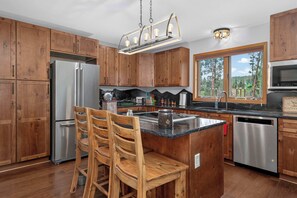 A kitchen with wooden cabinets, a stainless steel refrigerator, a built-in island with bar stools, and a window with a view of trees. - Charming 3 Bed Cabin| Lakes, Hiking, Hot Tub (Grand Lake)
