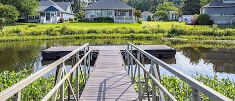 A wooden dock leading to a house with a porch, surrounded by lush greenery and a calm water body.
