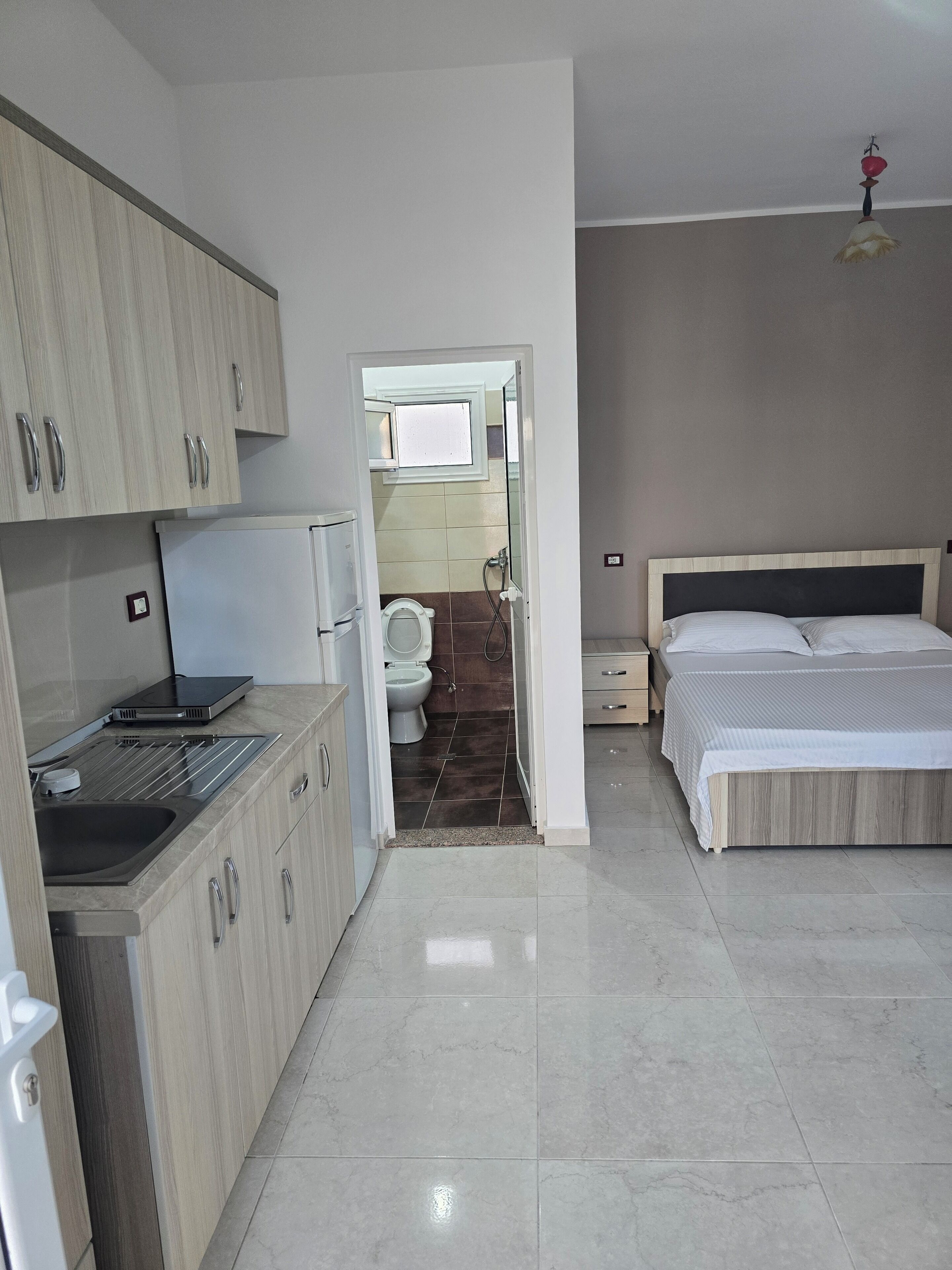 Habitación de hotel moderna con cocina, baño con inodoro y ducha, y cama con cabecero.