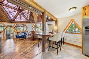 Ein geräumiger Raum mit hohen Decken, Holzbalken, einem Esstisch mit Stühlen und einem großen Fenster mit Blick auf Grünflächen.