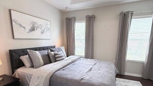Un dormitorio con cama, ventilador de techo, mesita de noche, alfombra y ventana con cortinas.