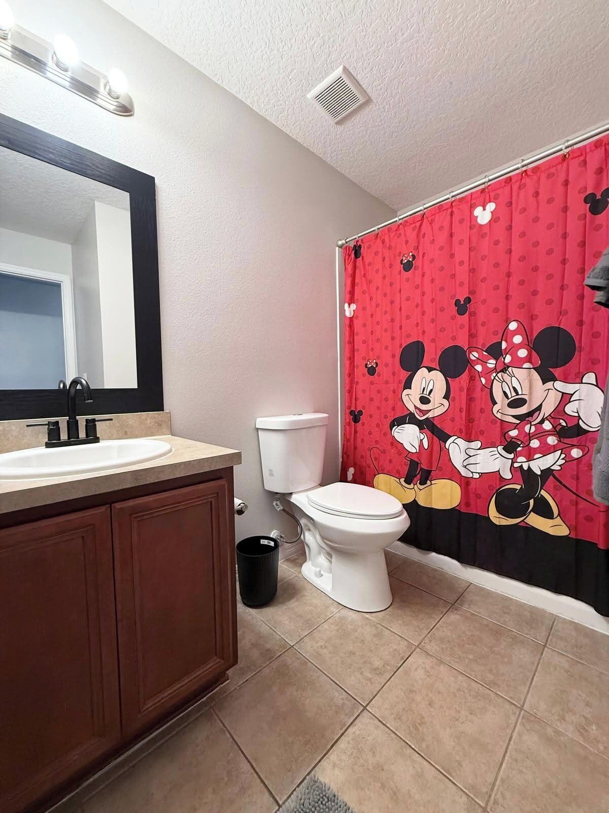 A bathroom with a toilet, sink, mirror, and Mickey Mouse themed shower curtain.