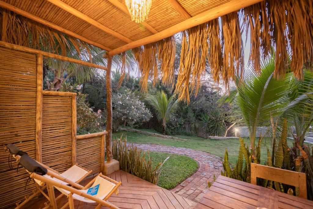 Un espacio de estar techado al aire libre con un banco de madera y una silla, rodeado por un jardín con palmeras y un sendero de ladrillo.