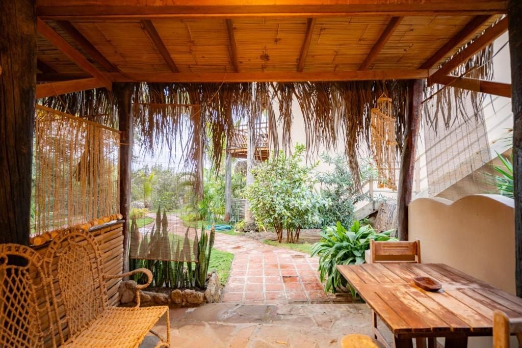 A covered outdoor seating area with a wooden bench and table, surrounded by plants and a stone pathway.