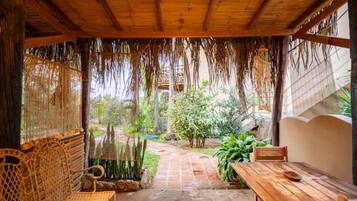 A covered outdoor seating area with a wooden bench and table, surrounded by plants and a stone pathway.